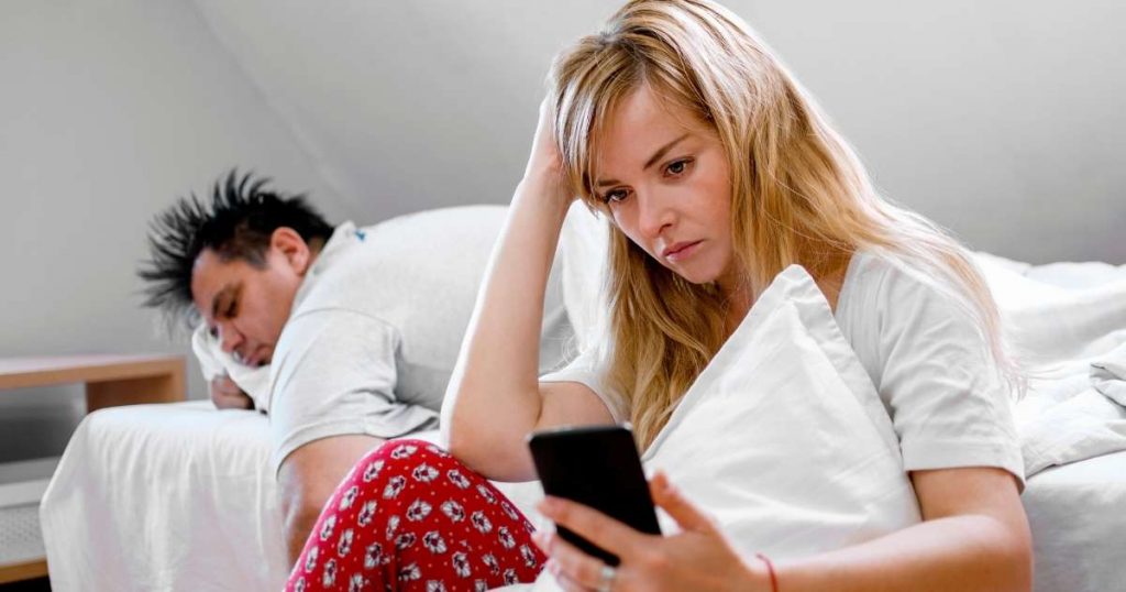 Wife using mobile phone hiding from husband who is sleeping in bed