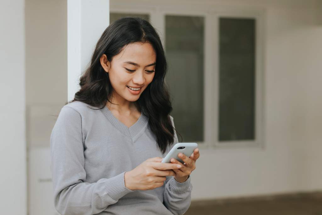 A young woman holding a phone and smiling joyfully, possibly while texting with the person she wants to make miss her.
