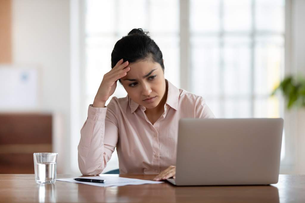 A person sitting at a desk, surrounded by scattered papers and looking frustrated while staring at a computer screen.